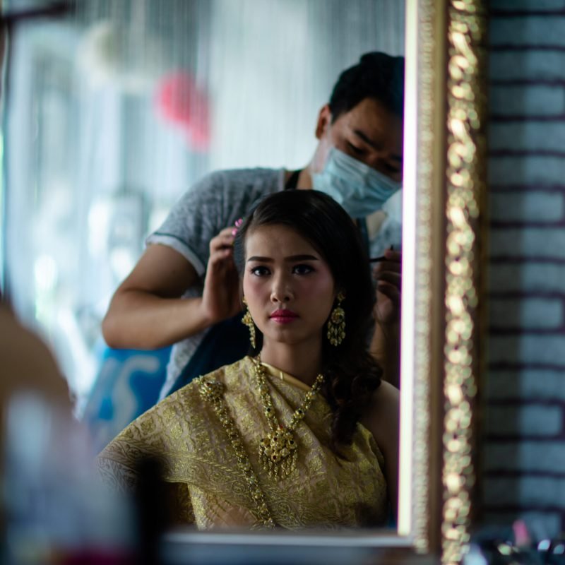 female hairdresser standing, face makeup and making hairstyle to cute lovely young woman in beauty salon